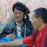 A long-haired nomad and a monk talking near the prayer flag frame.